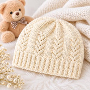 May include: A cream-colored knit beanie featuring a vertical cable stitch pattern and a ribbed brim. The hat is crafted from soft yarn and displays a consistent texture throughout, showcasing a classic design with a folded edge detail.