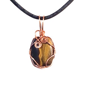 May include: A pendant featuring a polished, oval-shaped tiger's eye stone, wrapped in copper wire. The stone displays bands of brown and gold. The pendant hangs from a black cord, creating a simple, elegant necklace.