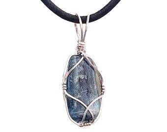 Puede incluir: Colgante de kianita azul de forma irregular, envuelto en alambre plateado, suspendido de un cordón negro. El alambre está intrincadamente tejido alrededor de la piedra. Un collar único.