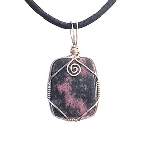 May include: A square pendant necklace featuring a dark stone with pink and black marbling, encased in a silver wire frame. The wirework includes a spiral design and is suspended from a black cord.