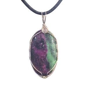 May include: Oval pendant with a polished ruby zoisite stone, showcasing a blend of deep red, green, and black hues. The stone is encased in a silver wire wrap and suspended from a black cord necklace.