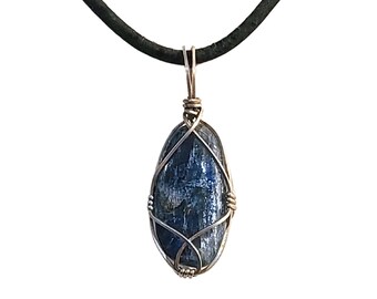 Puede incluir: Colgante de kianita azul con un alambre plateado, suspendido de un cordón negro. La piedra ovalada tiene una superficie pulida y está encerrada en un diseño decorativo de alambre. El colgante es una joya.