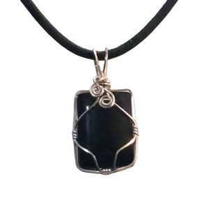 May include: A rectangular pendant with a dark, possibly black, stone is encased in silver-colored wire. The wire is intricately wrapped around the stone and forms a loop at the top, attached to a black cord necklace. The pendant hangs against a white background.