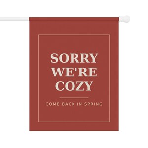 Puede incluir: Una bandera de jardín rectangular de color rojo terracota intenso con una fina línea de borde blanca. El texto SORRY WE'RE COZY está impreso en letras mayúsculas blancas grandes, seguido de una línea horizontal y el texto COME BACK IN SPRING.