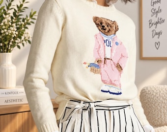May include: A cream-coloured long-sleeve knit jumper featuring a graphic of a teddy bear wearing a pink suit and blue scarf. The bear holds a small blue basket. The jumper has a crew neckline and is layered over a white collared shirt.