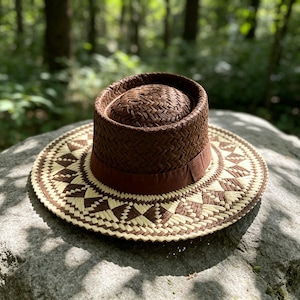 Puede incluir: Un sombrero de paja marrón oscuro con copa plana y ala ancha. El ala presenta un patrón geométrico en tejido de paja alternando marrón oscuro y crema. Una cinta de color marrón oscuro sólido rodea la base de la copa, creando un contraste.