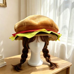 May include: A plush hat shaped like a hamburger, featuring a tan bun top with white embroidered sesame seeds, a red layer representing tomato, and a green layer representing lettuce, all resting on a white mannequin head with brown hair.