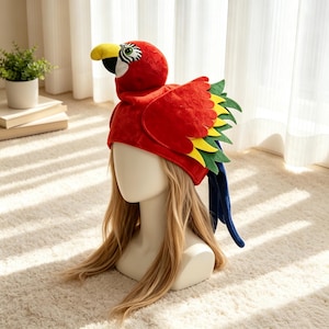 May include: A plush parrot hat featuring a bright red head with a yellow beak, white and black eye patches, and layered felt wings in red, yellow, and green. The hat includes long, dark blue tail feathers extending from the back of the head.
