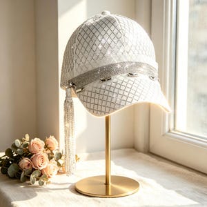 Op de afbeelding: Een witte baseballpet met een ruitpatroon van kleine zilveren strass-steentjes. De pet is voorzien van een brede, glinsterende zilveren band aan de voorzijde en een lange, bungelende zilveren kettingkwast die aan het zijpaneel is bevestigd.