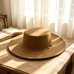Puede incluir: Un sombrero de fieltro color canela con copa plana y ala ancha. Una fina banda metálica circular de tono dorado rodea la base de la copa, con dos pequeñas cuentas esféricas doradas en la parte frontal donde se unen los extremos.