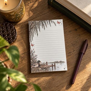 May include: A rectangular notepad with horizontal lines featuring a printed illustration of a mallard duck near a wooden dock, surrounded by willow branches, wildflowers, and small butterflies in shades of brown, green, and soft pink.