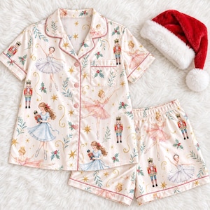 May include: A short-sleeve button-down pajama shirt and matching shorts set in a cream color with a repeating print of ballerinas, nutcrackers, holly leaves, and gold stars, accompanied by a red and white plush Santa hat with a white pom-pom.
