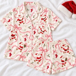 Può includere: Un set da pigiama composto da una camicia a maniche corte con bottoni e pantaloncini abbinati color crema, caratterizzato da un motivo ripetuto di Babbo Natale, calze rosse e nastri rosa con foglie di agrifoglio verdi e piccole stelle dorate.