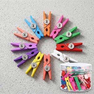 May include: A collection of twelve wooden clothespins in various colors including blue, orange, pink, green, red, white, yellow, and purple, arranged in a circular pattern, with a clear plastic jar filled with additional colorful clothespins.