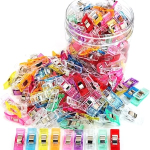Könnte beinhalten: Eine Sammlung von durchsichtigen Kunststoff-Nähklammern in verschiedenen leuchtenden Farben wie Rot, Blau, Gelb, Grün, Pink und Transparent. Die Klammern haben Metallfedern und befinden sich in einem durchsichtigen Kunststoffbehälter.