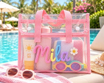 May include: A clear pink mesh tote bag featuring the name Mila in colorful raised script lettering. The bag contains a sunscreen bottle labeled SPF 50, a water bottle, a striped pouch, and yellow-rimmed goggles with a small flower patch.