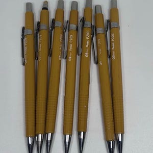 以下が含まれることがあります： 銀色の金属製クリップとペン先を備えた7本の黄色いシャープペンシル。各ペンシルには、本体に0.9mm Pentel P209という文字が印刷されています。ペン先付近にはリブ付きのグリップセクションがあり、上部には銀色のノック式メカニズムが付いています。