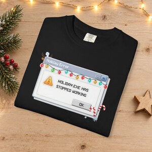 Può includere: Una t-shirt nera con una grafica pixelata di una finestra di errore del computer che riporta Holiday Error e HOLIDAY.EXE HAS STOPPED WORKING. Il design include un'icona di avviso gialla e una serie di luci natalizie colorate.