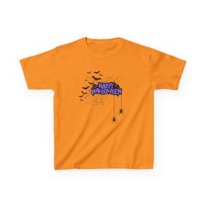 May include: A bright orange short-sleeved t-shirt featuring a black spiderweb graphic with silhouettes of flying bats. The text Happy Halloween is printed in purple with a black outline, accompanied by two black spiders hanging from the web.