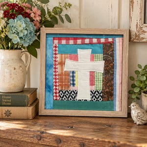 May include: A square framed wall art piece featuring a central white fabric cross appliqued onto a patchwork background. The quilt-style design incorporates various fabric strips in red, blue, green, and brown with plaid, floral, and geometric patterns.