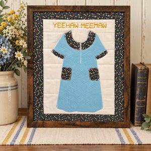May include: A framed fabric wall art piece featuring a light blue dress appliqué on a cream quilted background. The dress has a dark floral collar, sleeve cuffs, and pockets. The text YEEHAW MEEMAW is embroidered in yellow above the dress.