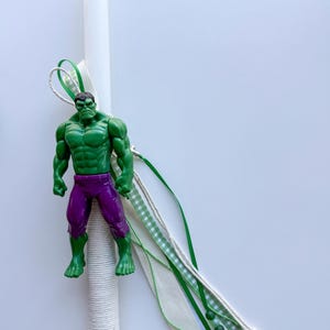 May include: A white candle decorated with a green and white ribbon and a Hulk figurine. The Hulk is green with purple pants. The candle is wrapped in white twine.