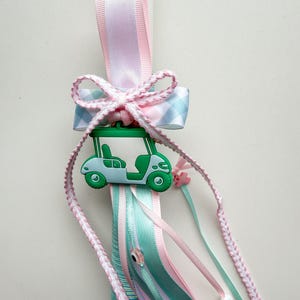 May include: A decorative candle with a golf cart charm. The candle features pink and blue ribbons, a gingham bow, and braided accents. The golf cart is green and blue. Beads and a small pink flower add detail.