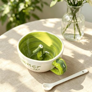 May include: A white ceramic mug with a green interior featuring a small, three-dimensional green crocodile figure at the bottom. The exterior has the word "summer" printed in green script, a green handle, and a matching white ceramic spoon.