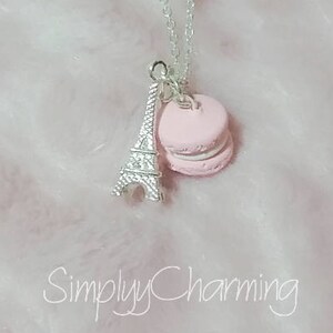 May include: A silver chain necklace with a pink macaron charm and a silver Eiffel Tower charm. The necklace is on a white fuzzy background. The text "Simply Charming" is visible in the bottom center of the image.
