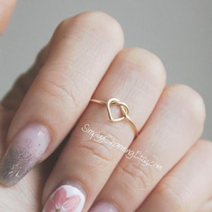 May include: A gold heart-shaped ring with a knot design. The ring is worn on the index finger of a hand with manicured nails.
