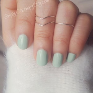 May include: A hand wearing three silver rings on the middle finger. The rings are simple and delicate. The nails are painted a light green colour.
