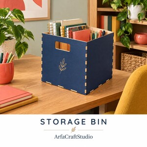 May include: A square navy blue storage bin with a cutout handle and a gold leaf line art design on the front. The bin features visible interlocking finger joints at the corners and is filled with various notebooks, journals, and pens.