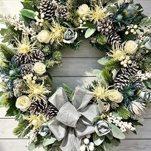 Peut inclure: Une couronne décorative composée de roses crème, de pommes de pin, de baies blanches et de feuillage vert. L'arrangement est agrémenté de fleurs jaune-vert épineuses et de boutons de rose bleu-gris, avec un grand nœud en tissu argenté scintillant.