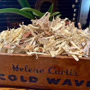 Può includere: Una scatola di legno piena di carta triturata. La scatola è marrone e ha le parole "Helene Curtis COLD WAVE" stampate in blu sul davanti.