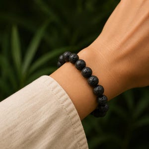 Puede incluir: Una pulsera compuesta por cuentas redondas de piedra de lava negra con una superficie porosa y texturizada. Las cuentas están ensartadas en un bucle continuo, creando una apariencia oscura y uniforme que contrasta con la manga de tela clara.