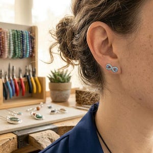 Puede incluir: Un pendiente de botón con forma de símbolo de infinito en tono plateado, adornado con pequeños cristales azul claro y un amuleto central de ojo turco en azul oscuro y blanco. El pendiente se lleva en el lóbulo y tiene acabado metálico.