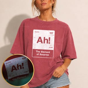 Puede incluir: Una camiseta de manga corta color granate con un gráfico rectangular blanco diseñado como un elemento de la tabla periódica. El gráfico muestra el texto Surprise, 117, Ah!, 14.994 y The Element of Surprise en un estilo de fuente desgastado.