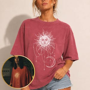 以下が含まれることがあります： 色あせた赤色の半袖Tシャツ。人間の顔をした太陽、垂れ下がるチェーン、三日月が描かれた白いグラフィックプリントが胸の中央に配置されています。クルーネックで、ゆったりとしたリラックス感のあるシルエットが特徴です。