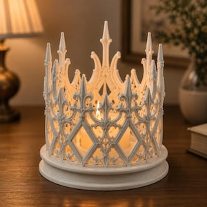 May include: A white candle holder featuring an intricate, openwork design reminiscent of gothic architecture with pointed spires and diamond-shaped cutouts. The circular base supports the structure, which glows from a lit candle inside.
