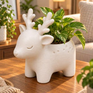 May include: A matte white ceramic planter shaped like a deer with closed eyes and textured antlers. The body features a series of small circular indentations along the back. A green and yellow variegated pothos plant grows from the open top.