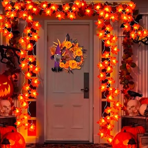 Pode incluir: Uma guirlanda de porta de Halloween com um gnomo de chapéu xadrez roxo, rosas laranja e pequenas abóboras, pendurada em uma porta branca emoldurada por dois fios verticais de luzes de folhas de bordo laranja e abóboras iluminadas.