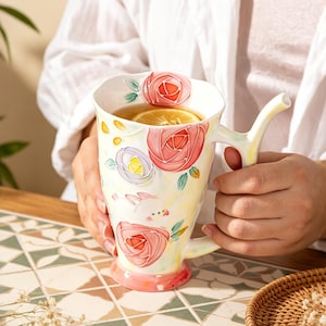 Könnte beinhalten: Ein Keramikbecher mit hellgelbem Hintergrund und erhabenen, bunten Blumenmotiven aus rosa und violetten Rosen, grünen Blättern und einem kleinen weißen Kaninchen. Der Becher hat einen astförmigen Henkel und enthält eine Zitronenscheibe.
