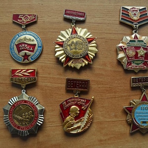 May include: A collection of six vintage Soviet-era military commemorative medals and badges. Each piece features intricate metalwork, enamel detailing in red, blue, and gold, and Cyrillic text. The designs include star shapes, soldier figures, and laurel motifs.