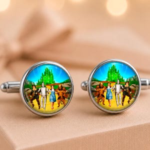May include: A pair of round silver-toned cufflinks featuring a colorful graphic print of the Wizard of Oz characters walking along a yellow brick road toward the Emerald City. The circular metal frames have a polished finish and standard toggle backs.