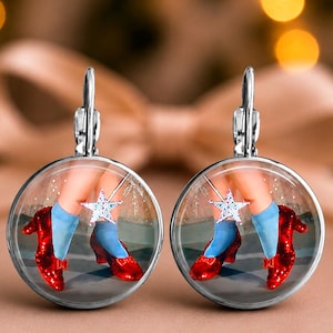 May include: A pair of round stainless steel earrings featuring a printed image of legs wearing light blue socks and shiny red slippers, with a small white star wand graphic. The text ruby slippers stainless earrings, great gift idea is visible.