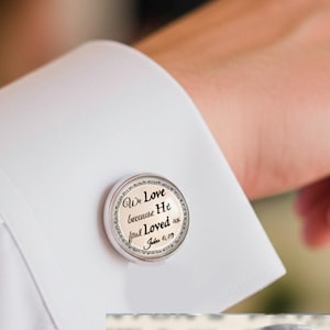 May include: Round silver-toned cufflinks featuring the text 'We Love because He first Loved us John 4:19' in black script on a cream background. The cufflinks have a decorative border and measure 18mm in diameter, shown on a white shirt cuff.