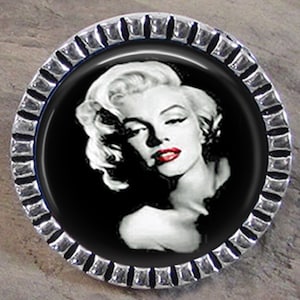 May include: A 25mm round clutchback pin featuring a black and white portrait of a woman with blonde hair and red lips. The pin has a silver-toned metal frame with a textured edge and a circular metal backing with a central clutch mechanism.