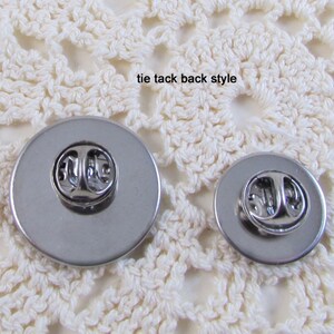 May include: Back view of two silver tie tacks with a pin and clutch fastening. The tie tacks are on a white crocheted surface.