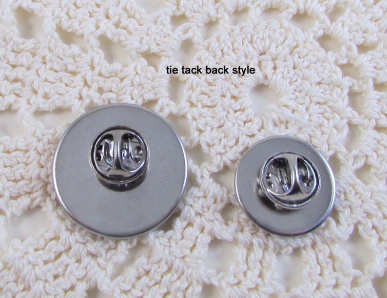May include: Back view of two silver tie tacks with a pin and clutch fastening. The tie tacks are on a white crocheted surface.