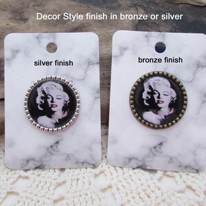 May include: Two round, silver and bronze, decorative brooches with a black and white image of Marilyn Monroe. The text "silver finish" and "bronze finish" is printed on the cards.